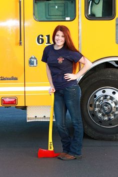 a woman standing in front of a yellow fire truck holding a shovel and smiling at the camera