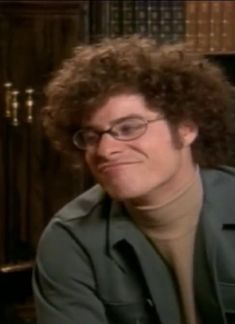 a man with curly hair and glasses sitting in front of a bookshelf