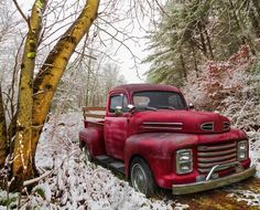 an old red truck parked in the snow next to a tree with no leaves on it