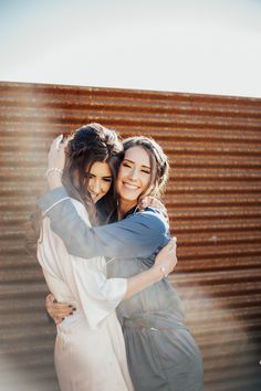 two women hugging each other in front of a corrugated wall with sunlight coming through the window