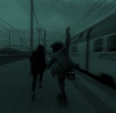 two people walking on the sidewalk next to a train track at night with their arms in the air