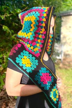 a woman wearing a colorful crocheted hood and black dress standing in front of trees