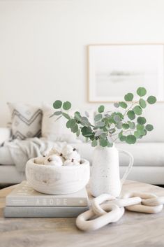 a table with a white vase filled with green plants on top of it next to a book