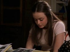 a woman sitting at a table reading a book with another person looking down on it