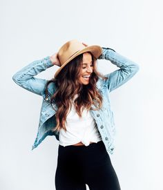 a woman in black pants and a white shirt is wearing a denim jacket with a hat on her head