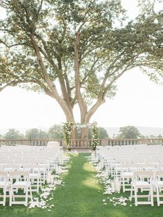 an outdoor ceremony set up with white chairs and flowers on the grass under a large tree