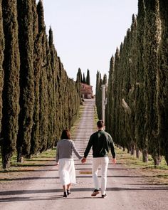 a man and woman holding hands while walking down a road lined with tall, thin trees