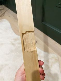 a hand holding a piece of wood in front of a green cabinet and white rug