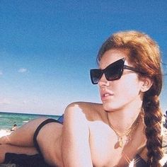 a beautiful young woman laying on top of a sandy beach next to the ocean wearing sunglasses