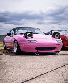 two pink sports cars parked next to each other on a road with cloudy skies in the background