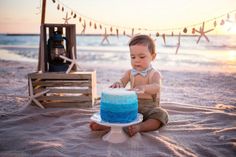 a baby boy sitting on the beach with a blue cake in front of him at sunset