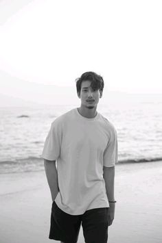 a young man standing on top of a beach next to the ocean with his hands in his pockets