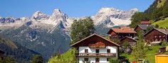 houses on the side of a mountain with snow capped mountains in the background and green grass below