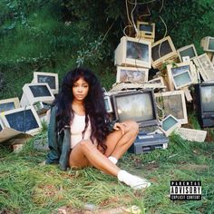 a woman sitting on the ground surrounded by old tvs and monitors in grass with trees behind her