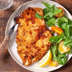 a white plate topped with chicken, salad and lemon wedges on top of a wooden table