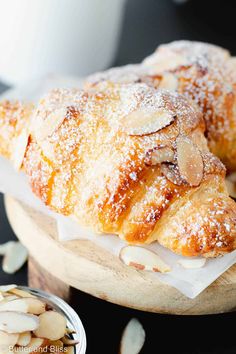 almond croissants with powdered sugar are on a wooden platter next to a bowl of almonds