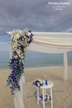 an outdoor wedding setup on the beach with white and purple flowers, blue accents and drapes