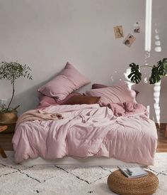 a bed with pink sheets and pillows in a room next to a potted plant