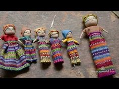 five dolls are lined up on the ground with knitting needles in their hands and one doll is wearing a multicolored knitted dress