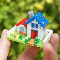 a hand holding a small toy house made out of plastic beads with a green tree in the background