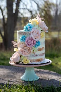 a multi - layered cake with flowers and butterflies sits on a pedestal in the grass