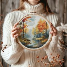 a woman holding up a cross - stitch autumn scene