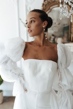 a woman wearing a white dress with ruffles on the shoulders and sleeves, standing in front of a mirror