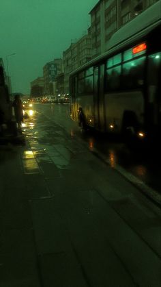 the bus is driving down the street at night time, with its lights on and it's raining