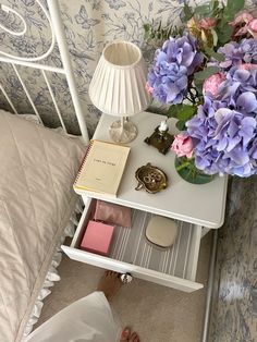 a white table topped with purple flowers next to a lamp and a book on top of a bed