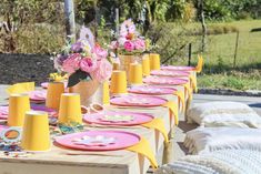 a long table is set with pink and yellow plates, vases filled with flowers