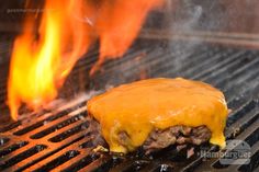 a hamburger covered in cheese on top of a bbq grill with flames behind it