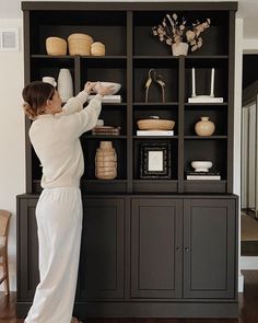 a woman is standing in front of a black bookcase with shelves and vases on it