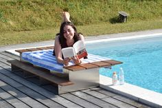 a woman laying on top of a wooden bench next to a pool reading a book