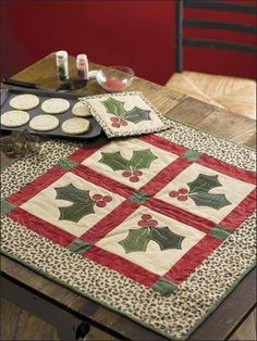 a quilted table topper with holly and cookies on it, sitting on a wooden table