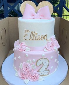 a pink and white two tiered cake with minnie mouse ears on top in a cardboard box