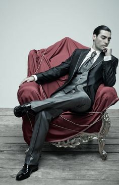 a man in a suit and tie sitting on a red chair with his hand under his chin