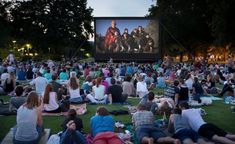 people are sitting on the grass watching a movie at night in front of a large screen