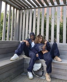 two people sitting on a wooden bench with their arms around each other and one person wearing a baseball cap