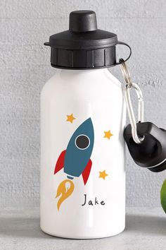 a white water bottle with a black lid and a rocket design on the side, sitting next to an apple