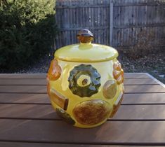 a yellow jar sitting on top of a wooden table next to a fence and bushes