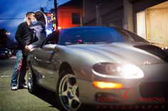 a man and woman kissing next to a silver sports car on the street at night