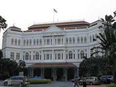 a large white building with cars parked in front of it and people walking on the sidewalk