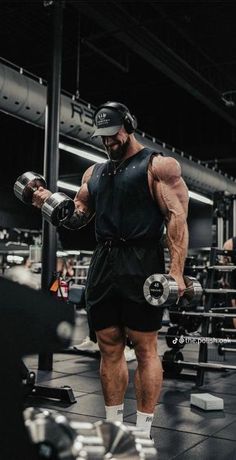 a man in black shirt and shorts holding two dumbs while standing in a gym