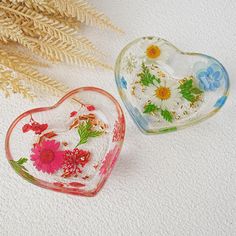 two glass heart shaped boxes sitting next to each other on a white surface with dried flowers