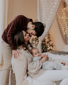 a man and woman kissing their baby on the forehead in front of a christmas tree