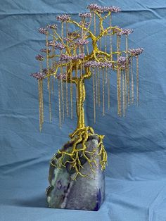 a tree made out of wire and beads sitting on top of a purple rock with blue background