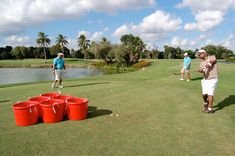 several people are playing golf on the grass near water and trees, with buckets in front of them