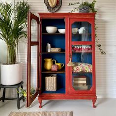 a red china cabinet with blue glass doors and shelves filled with dishes, vases and plants