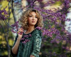 a woman with curly hair is standing in front of purple flowers and looking at the camera