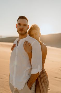 a man and woman standing in the sand at sunset, with one holding the other's back